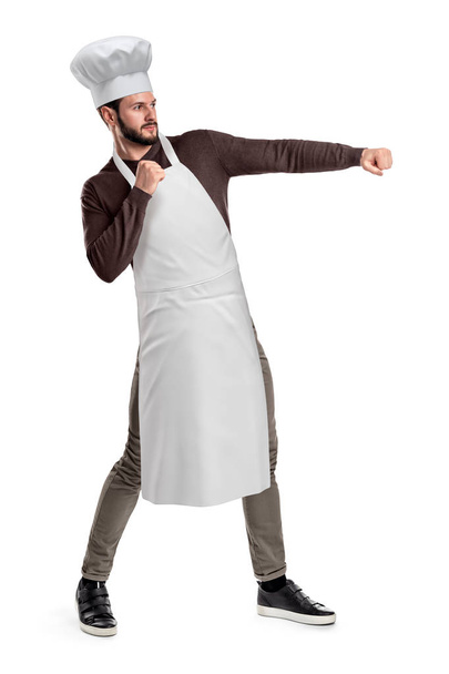 A young good-looking chef in a chef hat and apron posing with clenched fists and left hand held out as if about to hit something. - Foto, imagen