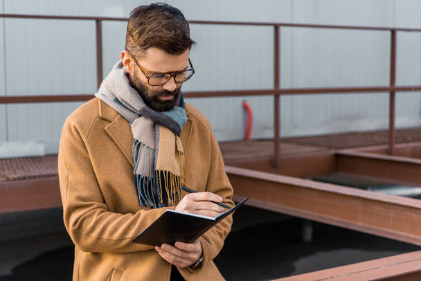 Bearded businessman standing in coat and writing in notebook  - Fotoğraf, Görsel