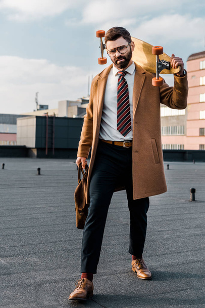 bearded businessman standing in formalwear and coat with penny board and bag in hands  - Фото, зображення
