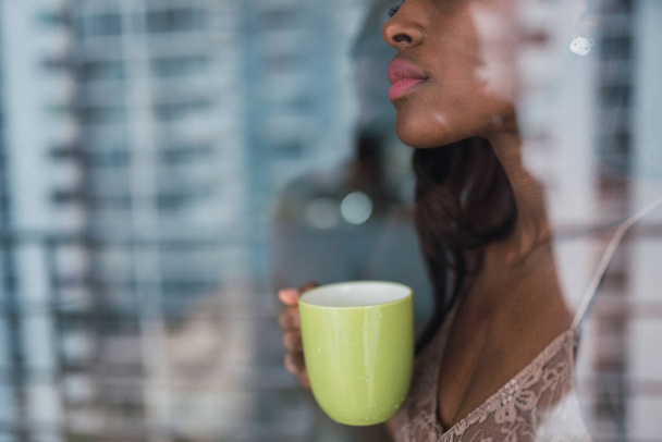 Shot through reflective window glass of alluring black woman with mug of coffee at home - Foto, afbeelding
