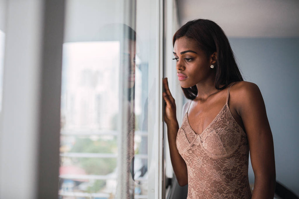 Sensual young African American woman in beige lace bodywear standing at window looking dreamily away - Fotografie, Obrázek