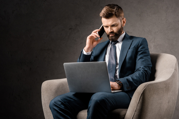 confident businessman sitting in armchair with laptop and talking on smartphone  - Foto, Imagem