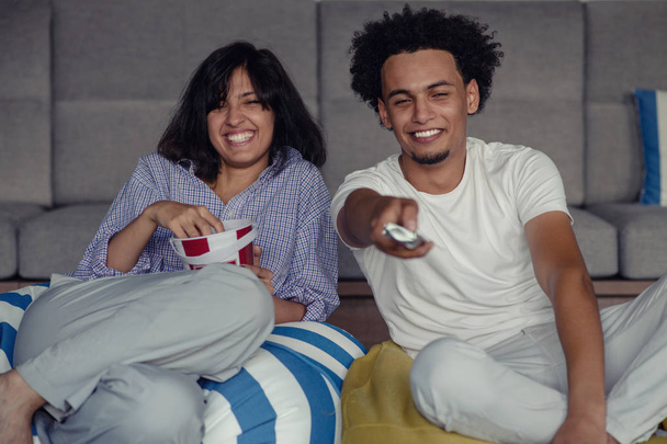 Image of smiling couple sitting on floor near sofa at home and looking aside while eating popcorn - Fotó, kép