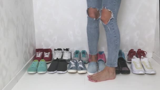 Una chica deportiva en jeans elige qué zapatillas de deporte para ir a entrenar. Varios pares de zapatos deportivos y piernas
. - Metraje, vídeo