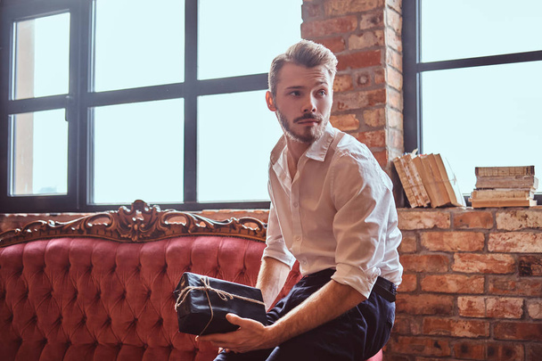 Un bel giovane uomo con una barba elegante e capelli elegantemente vestiti in possesso di una confezione regalo mentre seduto su un divano vintage rosso
. - Foto, immagini