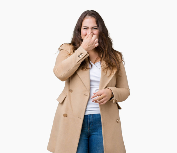 Beautiful plus size young woman wearing winter coat over isolated background smelling something stinky and disgusting, intolerable smell, holding breath with fingers on nose. Bad smells concept. - Photo, Image