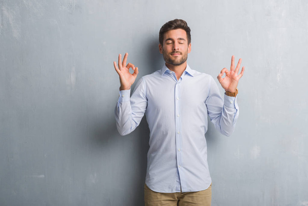 Handsome young business man over grey grunge wall wearing elegant shirt relax and smiling with eyes closed doing meditation gesture with fingers. Yoga concept. - Photo, Image