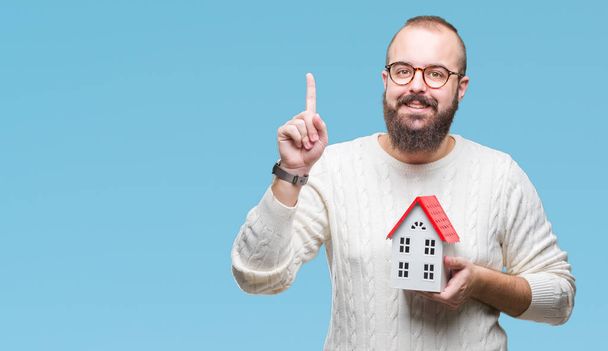 Young caucasian real state agent man holding house isolated background surprised with an idea or question pointing finger with happy face, number one - 写真・画像