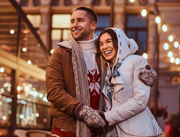Ένα νεαρό ζευγάρι ρομαντικό φοράει ζεστά ρούχα αγκάλιασμα δημοσια στο δρόμο το βράδυ στο χρόνο Χριστουγέννων, απολαμβάνοντας να ξοδεψουν το χρόνο μαζί. - Φωτογραφία, εικόνα