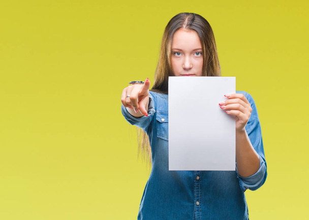 Young caucasian woman holding blank paper sheet over isolated background pointing with finger to the camera and to you, hand sign, positive and confident gesture from the front - Photo, Image
