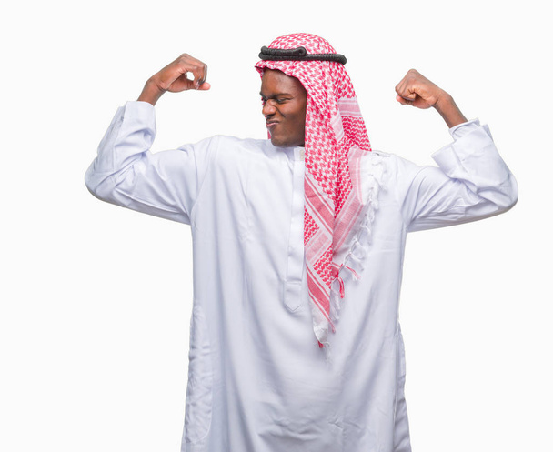 若いアラビアのアフリカ人の人は誇りに思って笑顔腕の筋肉を示す孤立した背景に伝統的なクーフィーヤを着てします。フィットネスの概念. - 写真・画像