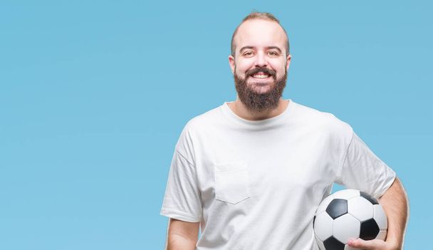 Young caucasian hipster man holding soccer football ball over isolated background with a happy face standing and smiling with a confident smile showing teeth - Photo, Image