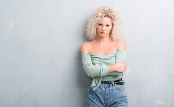 Young blonde woman with curly hair over grunge grey background skeptic and nervous, disapproving expression on face with crossed arms. Negative person. - Foto, immagini
