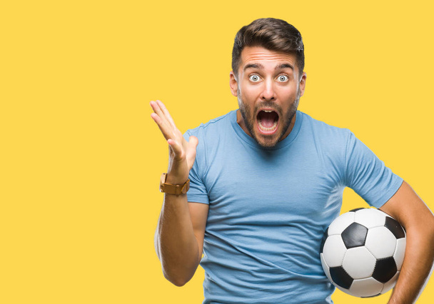 junger gutaussehender Mann hält Fußballball über isoliertem Hintergrund sehr glücklich und aufgeregt, Siegerausdruck feiert Sieg schreiend mit breitem Lächeln und erhobenen Händen - Foto, Bild