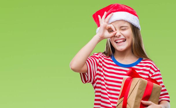 Молода красива дівчина в різдвяному капелюсі і тримає подарунок над ізольованим тлом з щасливим обличчям, посміхаючись, роблячи знак з рукою на око, дивлячись через пальці
 - Фото, зображення