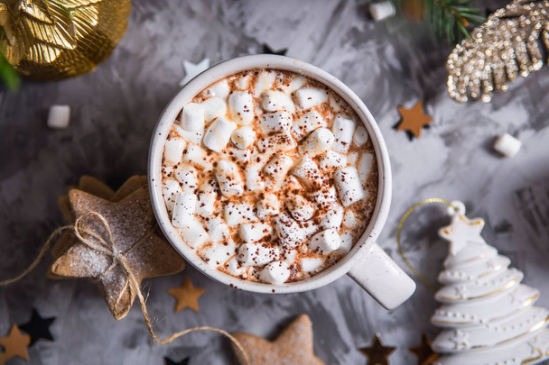 A large cup of cocoa with marshmallow sprinkled with cocoa powder stands on a gray table among Christmas decorations, fir branches, ginger cookies and shiny stars. Top view - Photo, Image