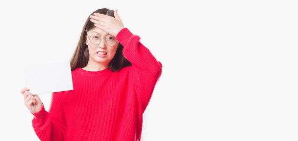 Jonge Chinese vrouw over geïsoleerde achtergrond met lege kaart benadrukt met de hand op het hoofd, geschokt met schaamte en verrassing gezicht, boos en gefrustreerd. Angst en boos voor fout. - Foto, afbeelding