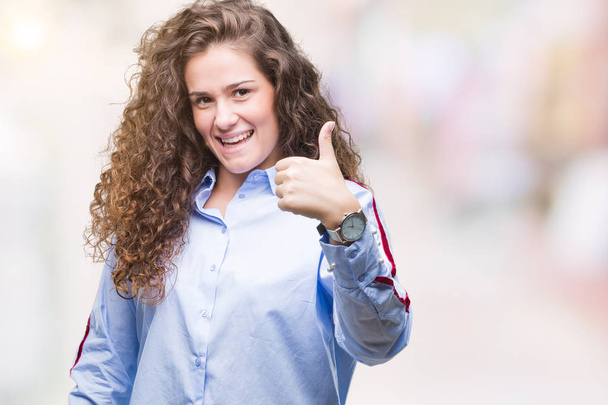 Beautiful brunette curly hair young girl wearing elgant look over isolated background doing happy thumbs up gesture with hand. Approving expression looking at the camera with showing success. - Photo, Image