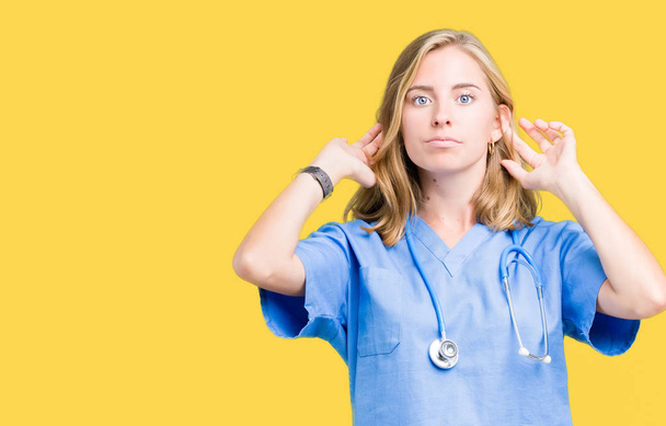 Красивая молодая доктор женщина в медицинской форме на изолированном фоне улыбаясь дергая за уши пальцами, смешной жест. Задача прослушивания
 - Фото, изображение