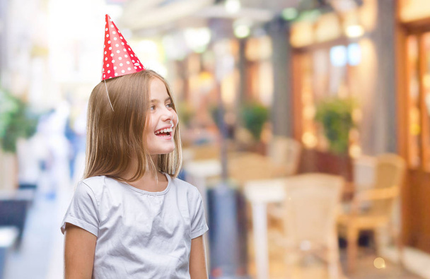 Young beautiful girl wearing birthday cap over isolated background looking away to side with smile on face, natural expression. Laughing confident. - Photo, Image