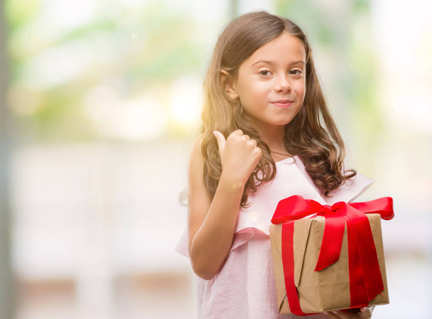 Brunette hispanique fille tenant un cadeau heureux avec un grand sourire faisant signe ok, pouce levé avec les doigts, excellent signe
 - Photo, image