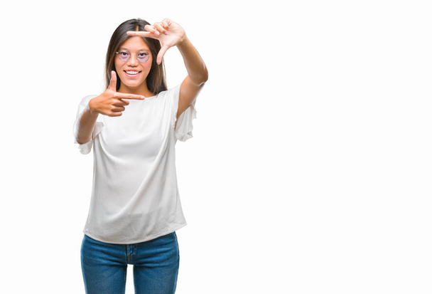 Jonge Aziatische vrouw bril over geïsoleerde achtergrond glimlachend maken frame met handen en vingers met blij gezicht. Creativiteit en fotografie concept. - Foto, afbeelding