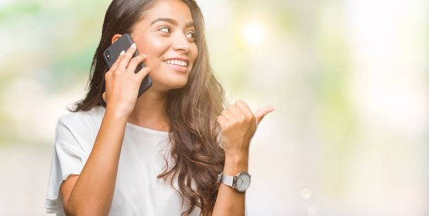 Young beautiful arab woman talking on the phone over isolated background pointing and showing with thumb up to the side with happy face smiling - Photo, Image