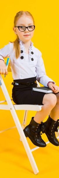 Girl with red hair on a yellow background. A charming girl in transparent glasses sits on a white chair and makes notes in a notebook. - 写真・画像