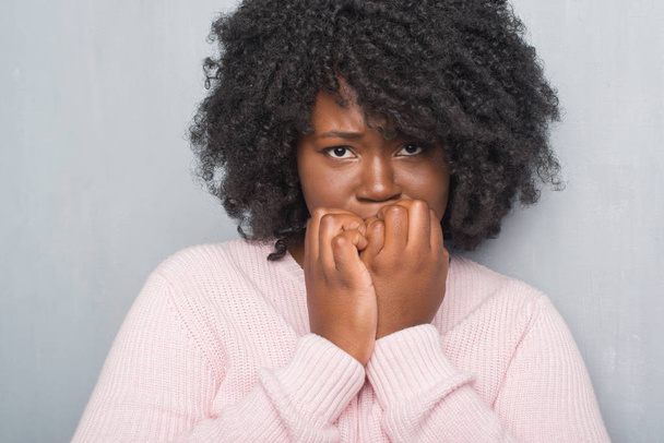 若いアフリカ系アメリカ人に加えて不安や両手の爪を噛んで口の中で緊張を見て冬のセーターを着ているグレーのグランジの壁を越えてサイズ女性。不安の問題. - 写真・画像