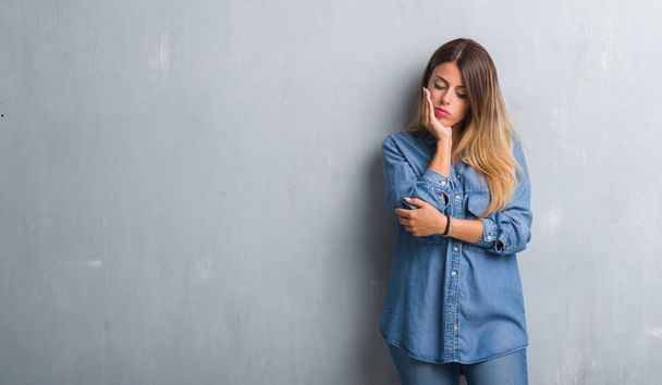 Молодая взрослая женщина за гранж серой стеной в джинсовой одежде думает устало и скучно с депрессией проблемы со скрещенными руками
. - Фото, изображение