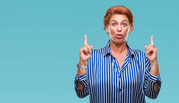 Atrractive senior caucasian redhead woman over isolated background amazed and surprised looking up and pointing with fingers and raised arms. - 写真・画像