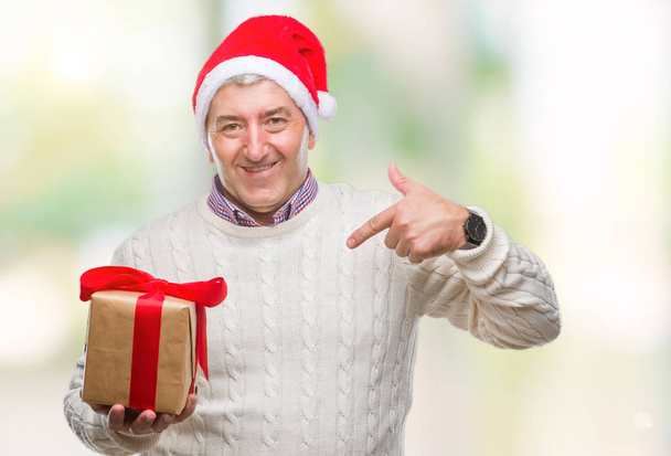 Hombre mayor guapo usando sombrero de Navidad y sosteniendo el regalo sobre un fondo aislado con la cara sorpresa apuntando con el dedo hacia sí mismo
 - Foto, imagen