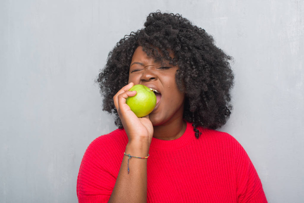 Jeune femme afro-américaine sur le mur gris grunge manger pomme verte avec une expression confiante sur la pensée intelligente visage grave
 - Photo, image