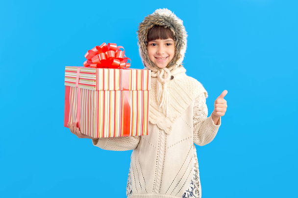Little girl in winter clothes with a big gift box in hands on a blue background. Show thumb up. Christmas holiday gifts. - 写真・画像