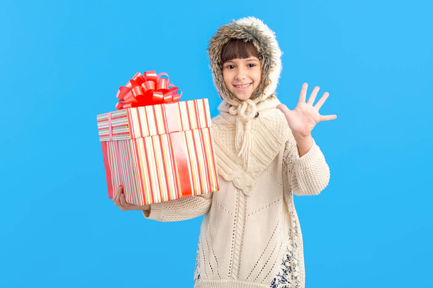 Little girl in winter clothes with a big gift box in hands on a blue background. Show five fingers. Christmas holiday gifts. - Photo, Image