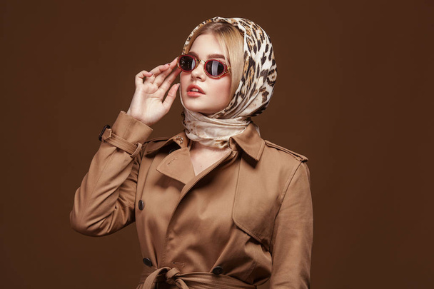 Fashion portrait of young stylish woman in trendy trench coat, leopard print headdress and fashion sunglasses over beige background. - Foto, afbeelding