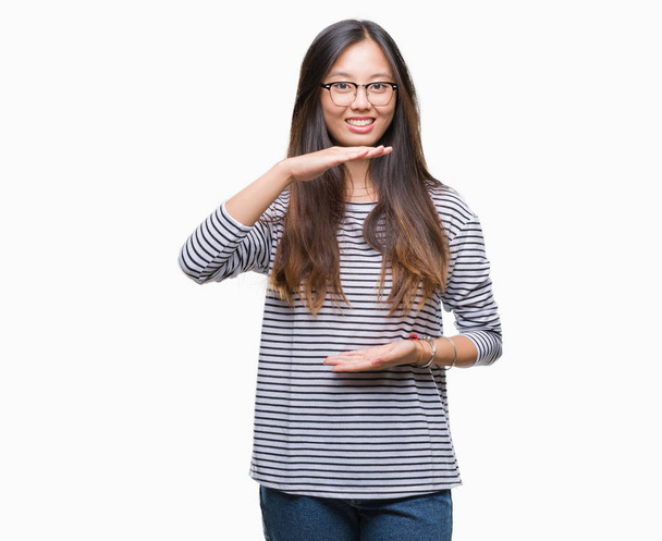 junge asiatische Frau mit Brille über isoliertem Hintergrund gestikuliert mit Händen, die große und große Zeichen zeigen, Maßsymbol. Lächelnd in die Kamera blickend. Messkonzept. - Foto, Bild