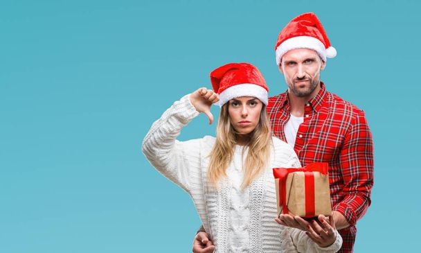 Young couple in love wearing christmas hat and holding present over isolated background with angry face, negative sign showing dislike with thumbs down, rejection concept - Photo, Image