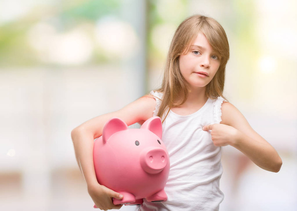 Young blonde toddler holding piggy bank with surprise face pointing finger to himself - Photo, Image