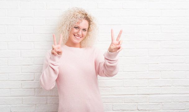 Sarışın genç kadın parmaklarını zafer işareti yaparken gösterilen kamera seyir gülümseyen beyaz tuğla duvarın üzerinden kıvırcık saçlı. İki numaralı. - Fotoğraf, Görsel