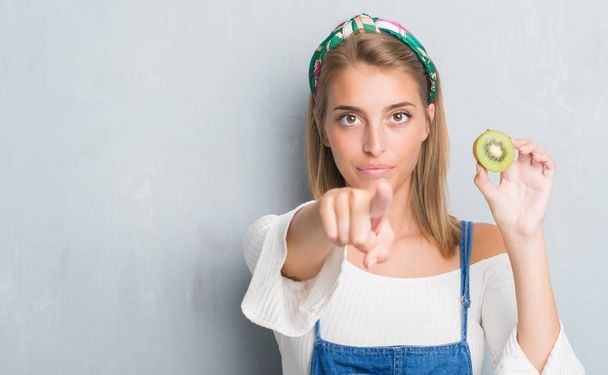 Gyönyörű fiatal nő eszik friss kiwi ujjal mutatva, hogy a kamera és az Ön grunge szürke falon, kéz jel, pozitív és magabiztos gesztus, az első - Fotó, kép