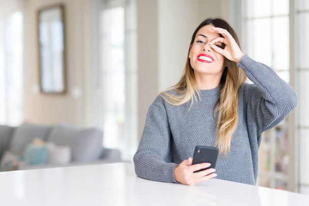 Młoda kobieta piękny szczęśliwy twarz uśmiechający się robi ok znak ręką na oko patrząc przez palce za pomocą smartfona w domu - Zdjęcie, obraz