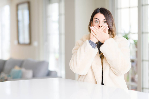 Mooie jongedame dragen winterjas bont thuis geschokt bedekking mond met handen voor fout. Geheime concept. - Foto, afbeelding
