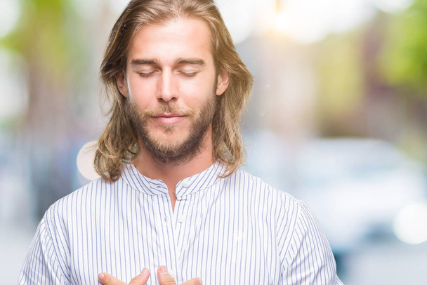 Young handsome man with long hair over isolated background smiling with hands on chest with closed eyes and grateful gesture on face. Health concept. - Photo, image