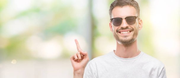 Jeune homme adulte tatoué portant des lunettes de soleil très heureux pointant avec la main et le doigt sur le côté
 - Photo, image