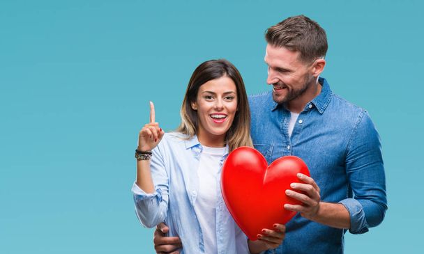 Young couple in love holding red heart over isolated background surprised with an idea or question pointing finger with happy face, number one - Fotografie, Obrázek