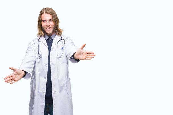 Young handsome doctor man with long hair over isolated background looking at the camera smiling with open arms for hug. Cheerful expression embracing happiness. - Photo, Image
