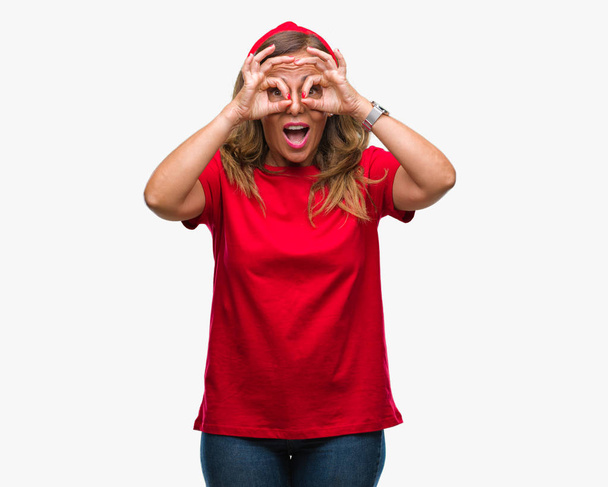 Středního věku starší žena, hispánský izolované pozadí dělá ok gesto jako dalekohled držet jazyk, oči při pohledu přes prsty. Šílený výraz. - Fotografie, Obrázek