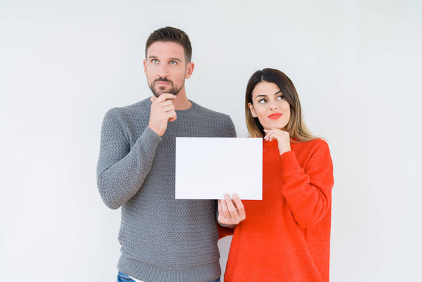 Young couple holding blank sheet paper over isolated background serious face thinking about question, very confused idea - Fotoğraf, Görsel
