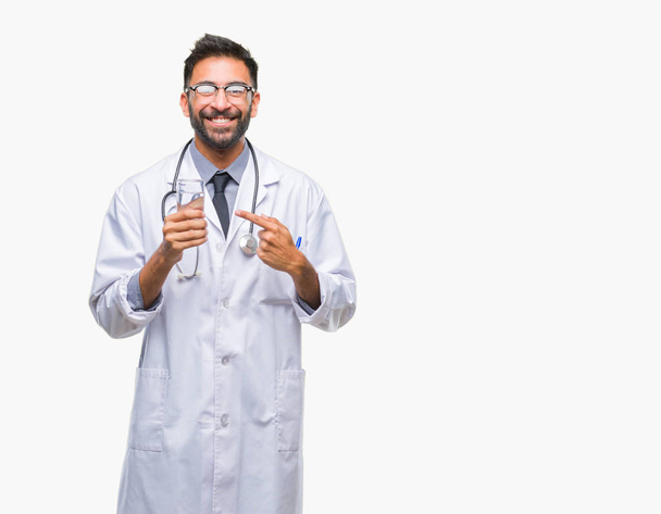 Дорослий іспаномовний лікар чоловік п'є склянку води над ізольованим тлом дуже щасливий вказуючи рукою і пальцем
 - Фото, зображення